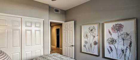 A bedroom with a bed, a ceiling fan, two framed floral artworks, and a door leading to another room.