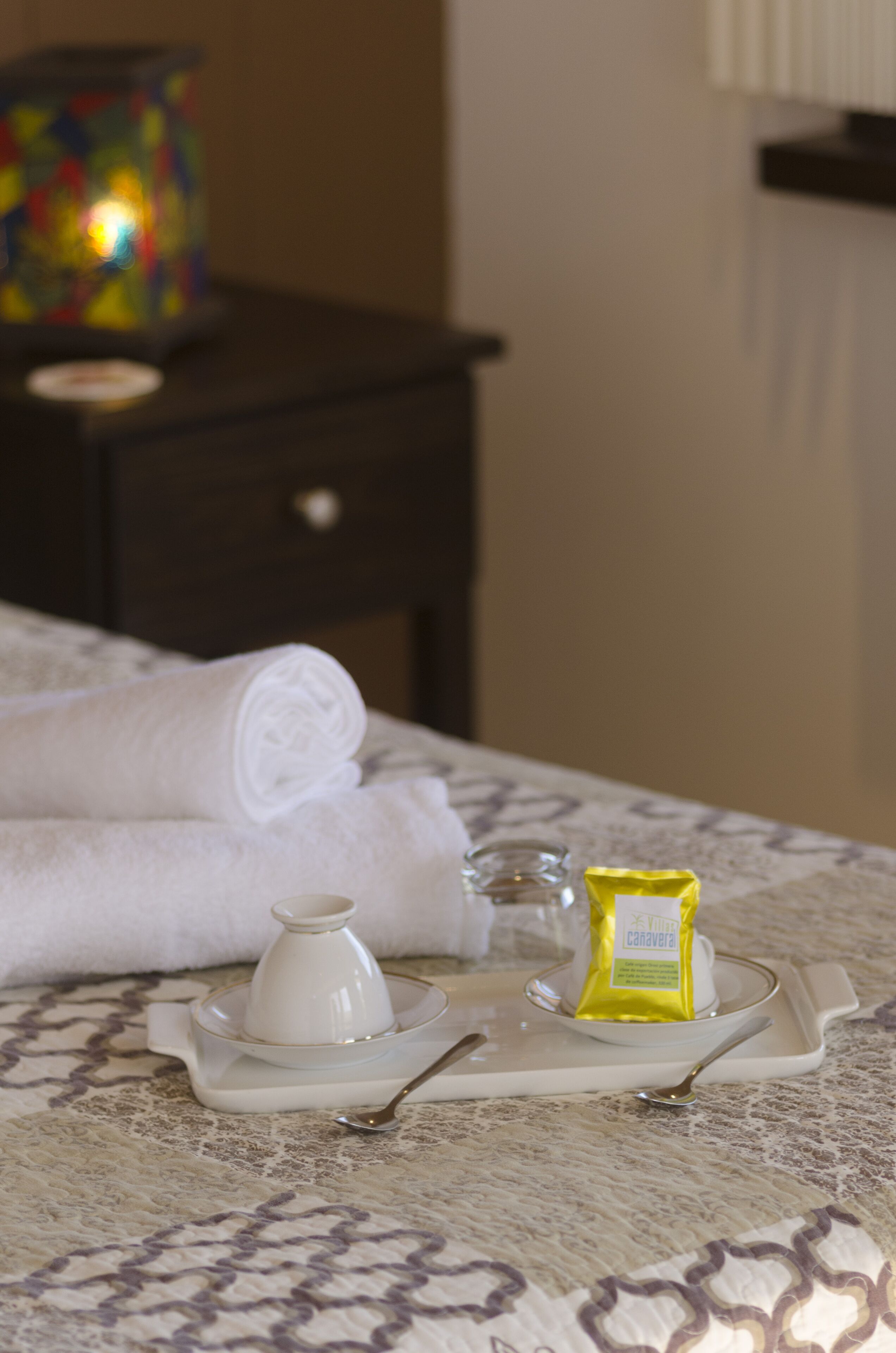 A neatly made bed with a tray containing a white cup, a small glass container, and a yellow packet, likely containing a snack or toiletry item.