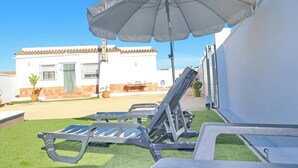 Een terras met een wit gebouw op de achtergrond, een grote parasol en twee lounge fauteuils.