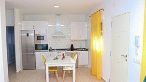 Een moderne keuken met witte kastjes, een roestvrijstalen koelkast, een magnetron en een spoelbak. Er staat een eettafel met gele stoelen en een witte tafelkleed.