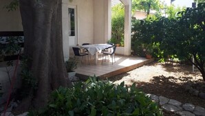 A covered outdoor seating area with a table and chairs, surrounded by plants and a tree.