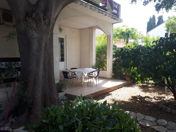A covered outdoor seating area with a table and chairs, surrounded by plants and a tree.