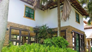 A building with a stone base and white upper walls, featuring green-framed windows and doors, surrounded by lush greenery. - OYO Pousada do Wagner, Rio das Ostras (Rio das Ostras)