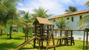 A wooden play structure in a grassy area with palm trees and a building with a tiled roof in the background. - OYO Pousada do Wagner, Rio das Ostras (Rio das Ostras)