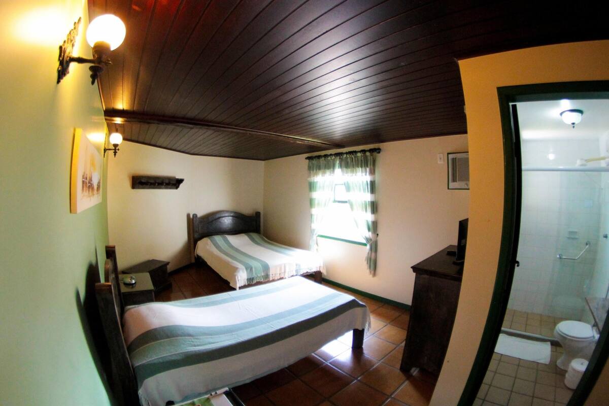 Ein Schlafzimmer mit Bett, Nachttischen, einem Fenster mit Vorhängen, einer kleinen Kommode und einem Badezimmer mit Toilette und Dusche.