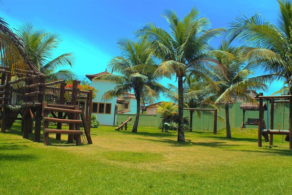 Un espacio al aire libre tropical con palmeras, una estructura de juego de madera y una casa al fondo.