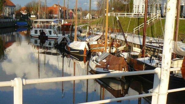 A marina with various boats docked, a white railing in the foreground, and buildings in the background. - Vacation home Ambiente, Germany (Wittmund)