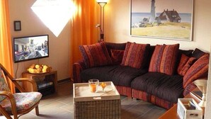 A cozy living room with a dark-colored sofa, a wicker coffee table, a television, and a painting of a lighthouse. - Vacation home Ambiente, Germany (Wittmund)
