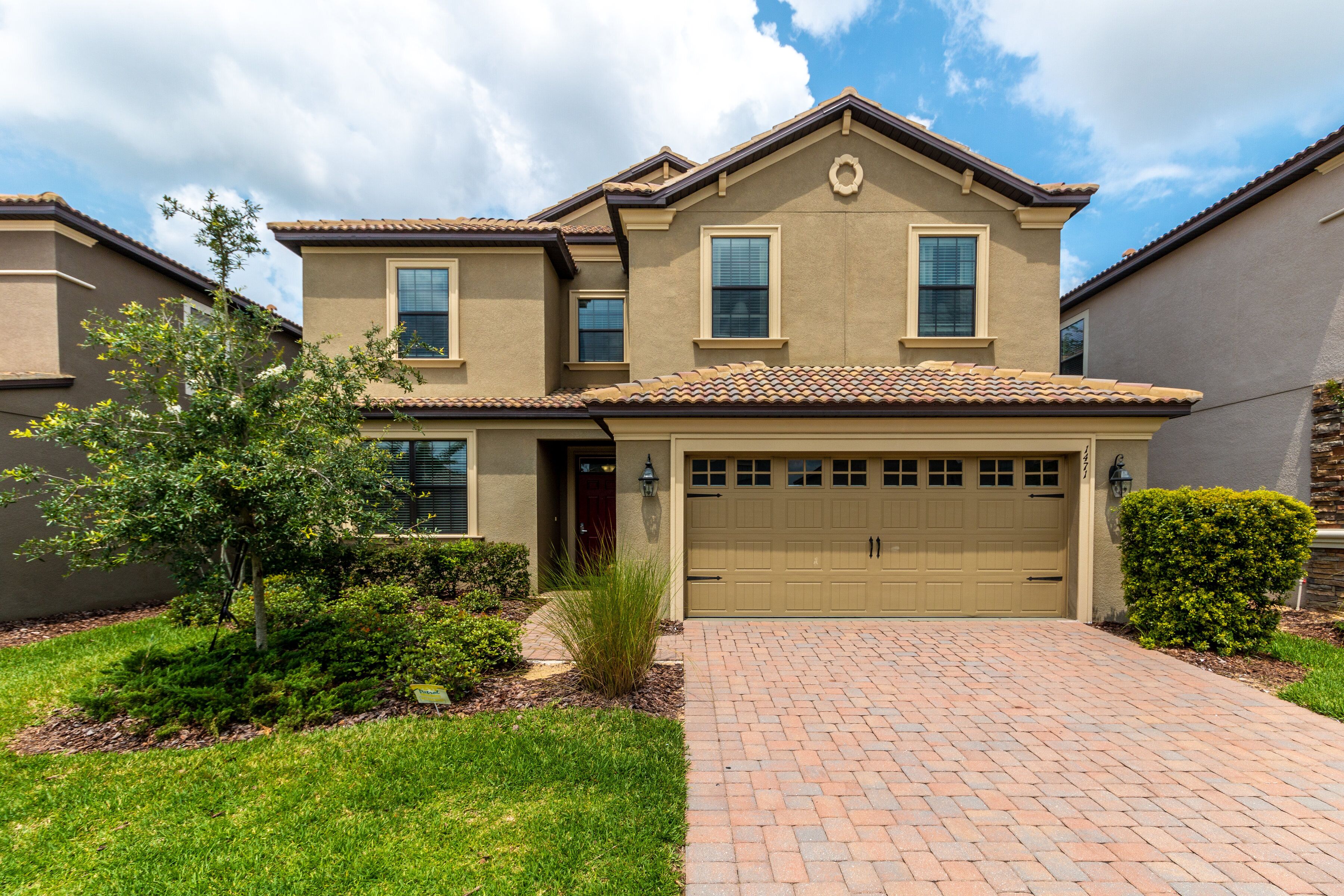 A two-story house with a beige exterior, a garage, and a brick driveway.