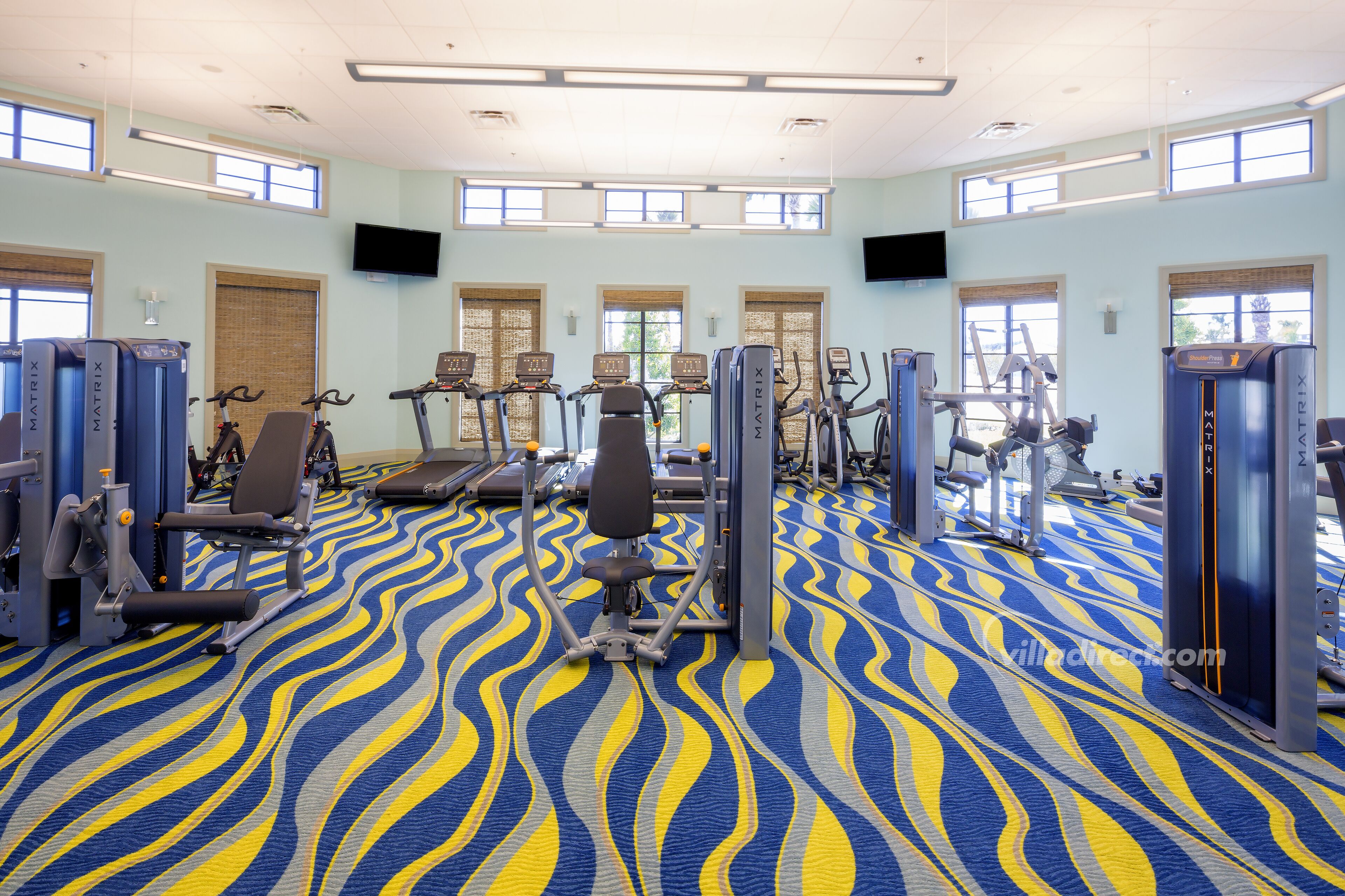 Ein Fitnessraum mit Trainingsgeräten, großen Fenstern und einem gemusterten Teppich.