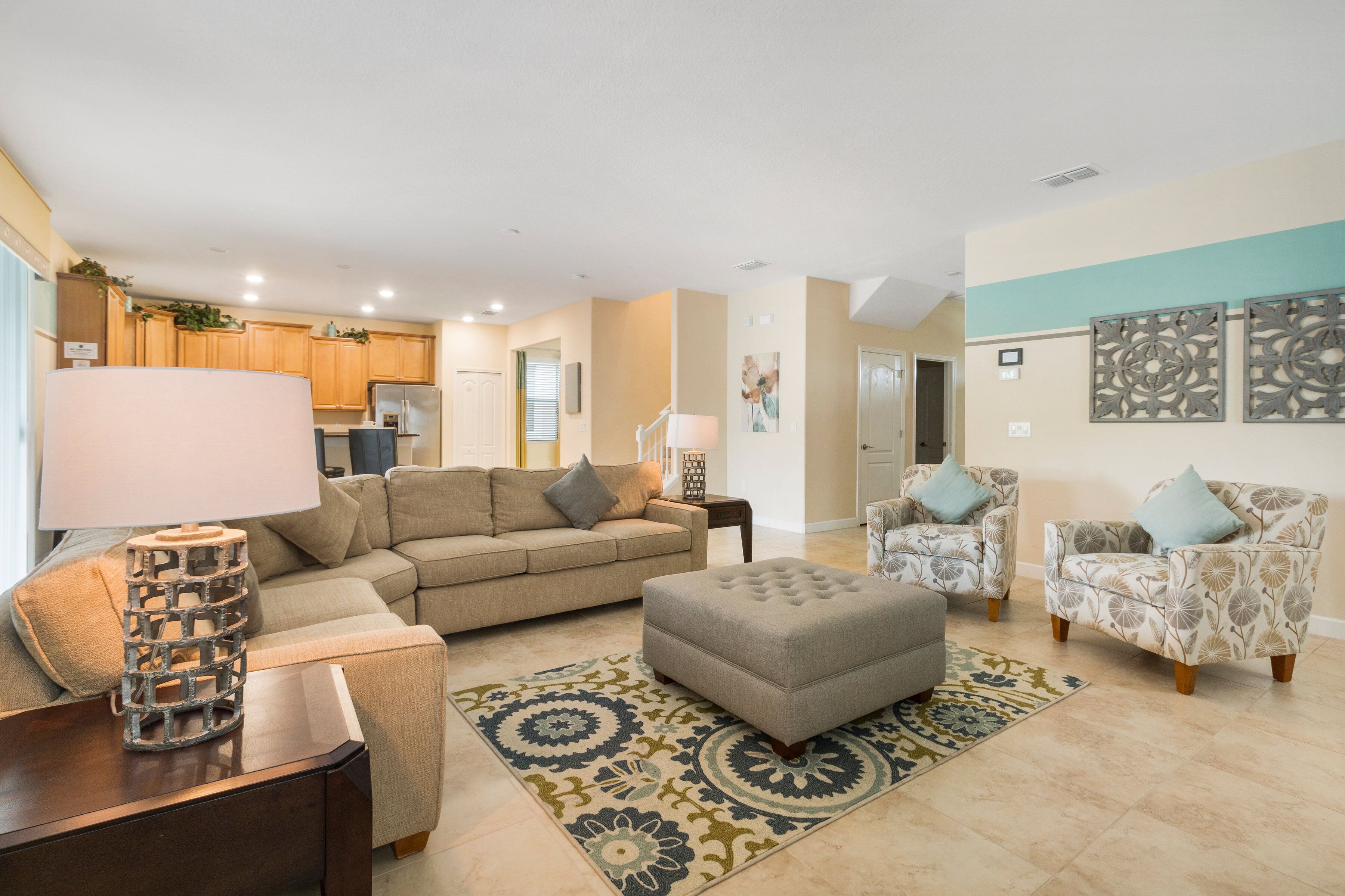 A modern living room with a sofa, ottoman, armchairs, and a coffee table. There is a kitchen in the background with wooden cabinets and a refrigerator.