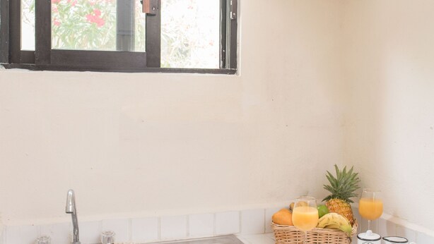A kitchen with a stainless steel sink, wooden cabinets, a window with a view of greenery, and a fruit basket with drinks on the counter. - SIMI-HA-2BR-TULUM... (TULUM)