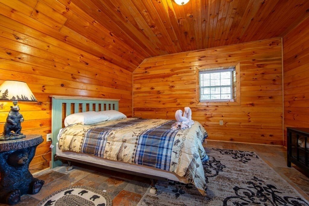 A wooden cabin bedroom with a bed, a window, a lamp, and a bear statue.