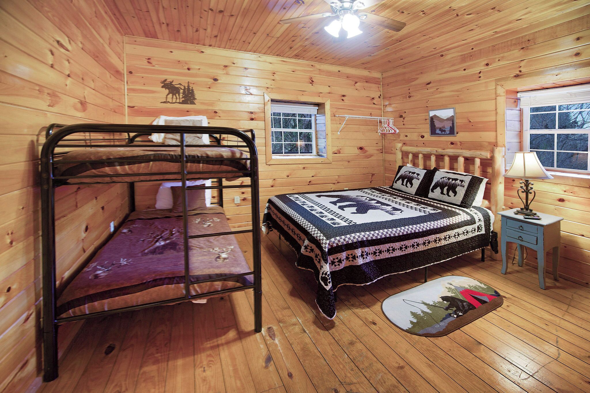 A wooden cabin bedroom with bunk beds, a bed with a patterned blanket, a small window, a lamp, and a picture on the wall.