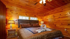 A wooden-paneled bedroom with a large bed, two windows, a nightstand, and a ceiling fan. -  Smoky Mtn Hideaway - 7BR, Hot Tub +Movie Theatre + Darts+ Firepit+ Pets ok (Sevierville)