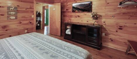 A wooden cabin room with a bed, a fireplace, and a door leading to another room.