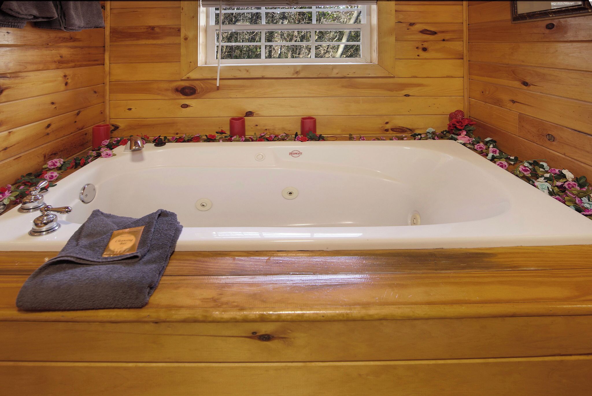A wooden cabin bathroom with a large bathtub, a window, and a towel on the floor.