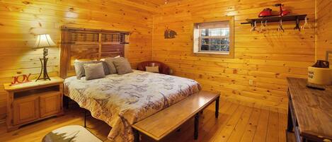 A cozy cabin-style room with a wooden floor, a bed with a floral bedspread, a bench, a nightstand with a lamp, and a shelf with a red kettle.