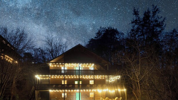 A two-story wooden cabin with string lights along the railings and a green door, surrounded by trees. -  Smoky Mtn Hideaway - 7BR, Hot Tub +Movie Theatre + Darts+ Firepit+ Pets ok (Sevierville)