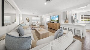 A spacious living room with a large sofa, a wooden coffee table, and a TV. There is a kitchen area with white cabinets and a bar counter.