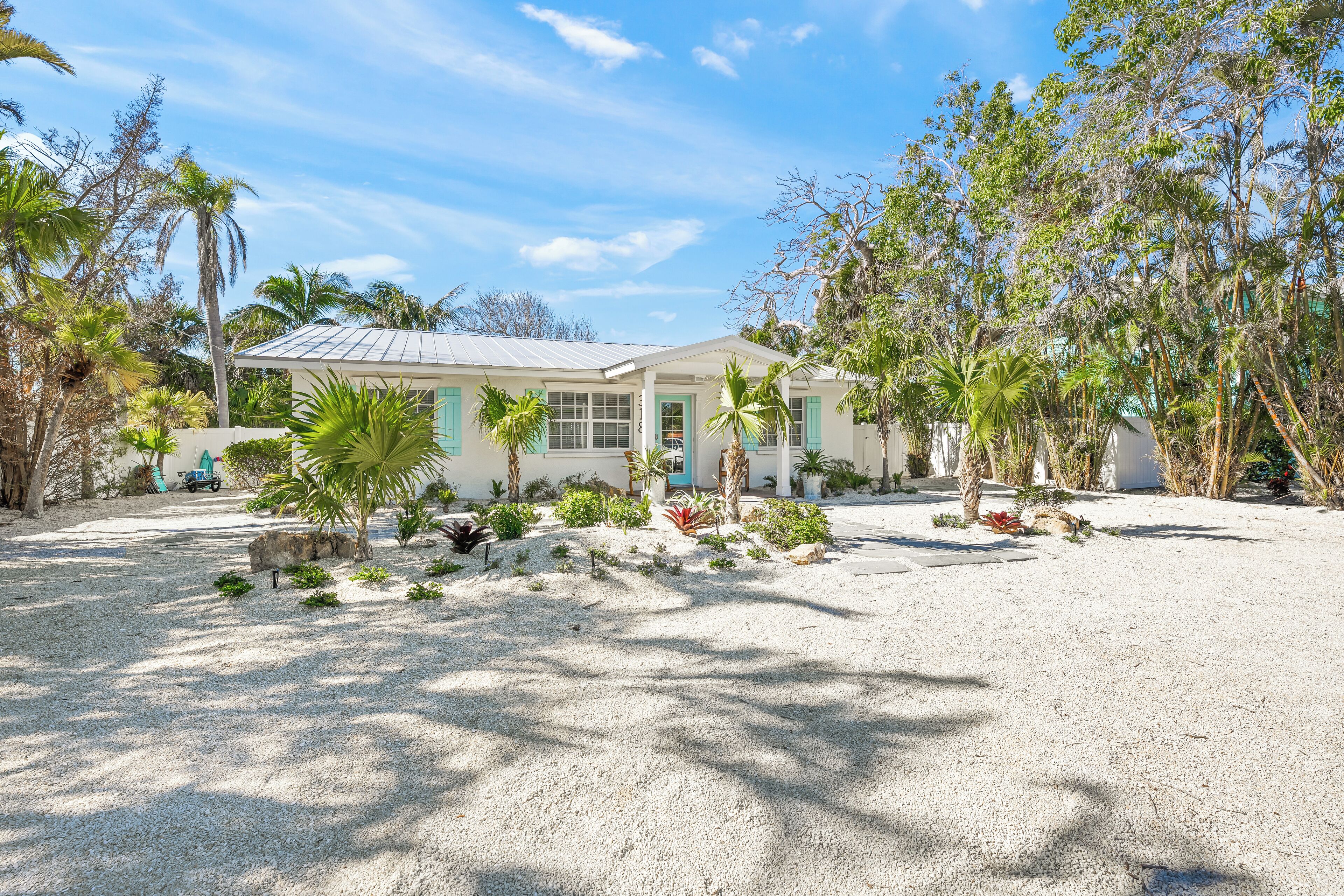 A house with a white exterior, a green door, and a gravel driveway surrounded by tropical plants.