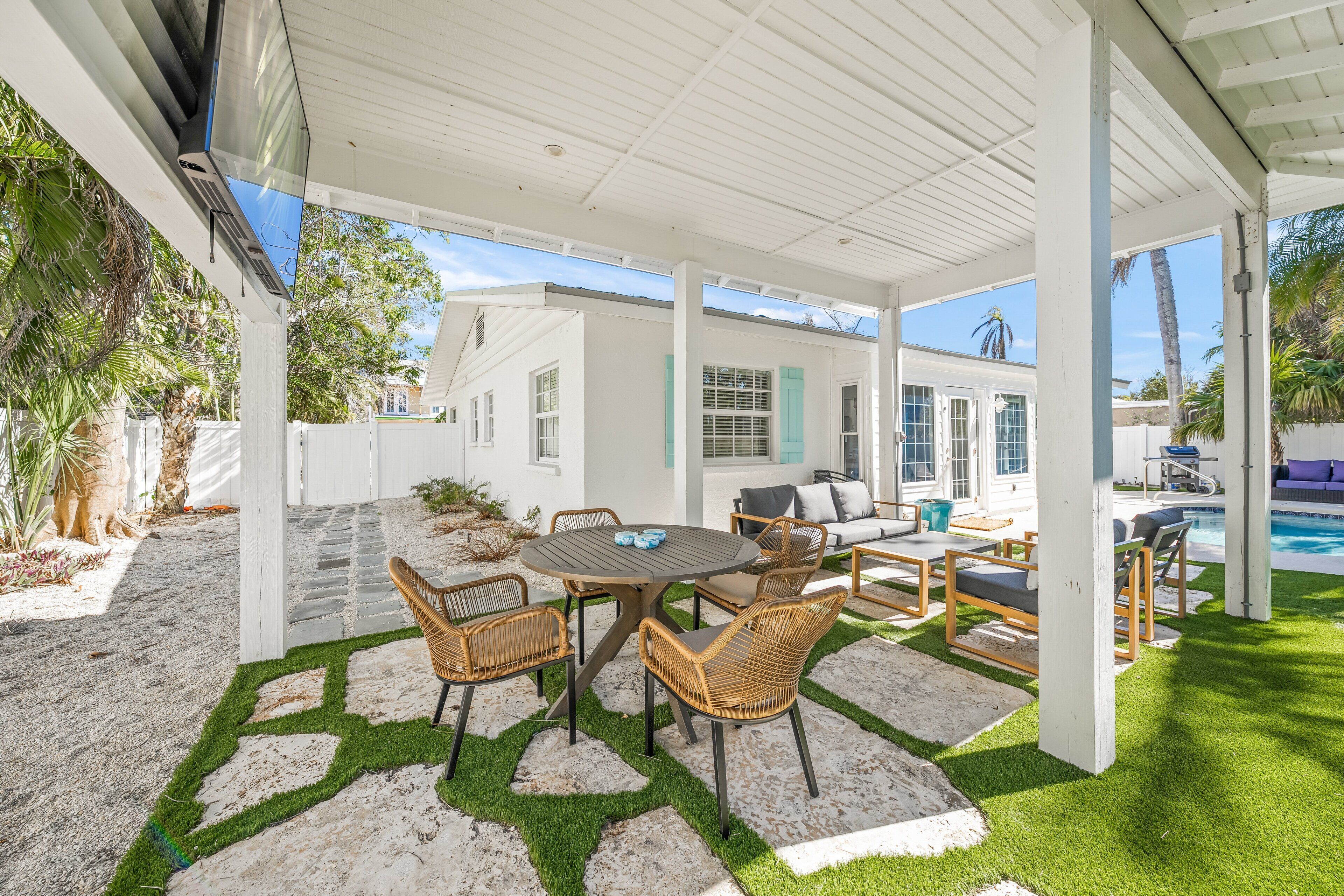 Une terrasse extérieure couverte, avec une table et des chaises, une piscine et un bâtiment blanc en arrière-plan.