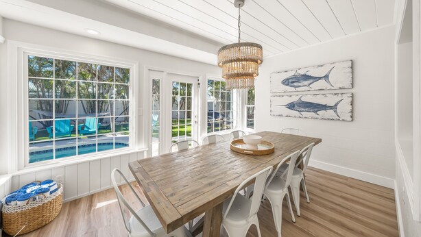 A dining area with a wooden table, white chairs, a chandelier, and a view of a pool and outdoor seating.