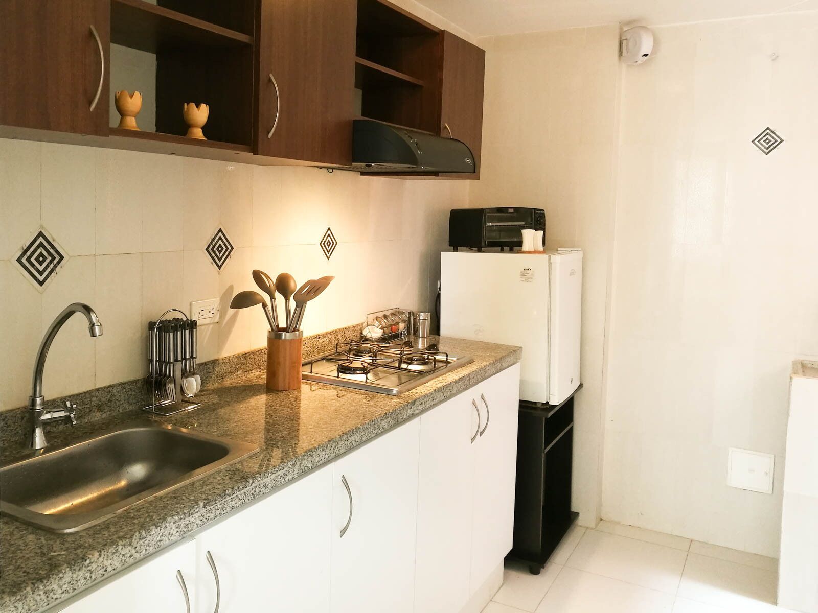 Una cocina con encimeras de granito, pileta de acero inoxidable y gabinetes blancos. Cuenta con una estufa a gas, un tostador negro y un refrigerador blanco.
