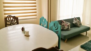 Una habitación con una mesa de comedor redonda, dos sillas, un sofá con cojines estampados y una ventana con persianas.