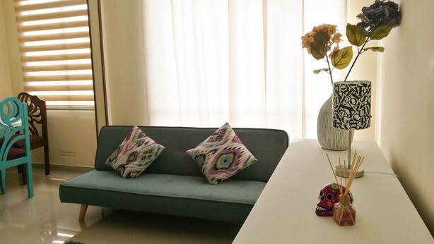 Una sala de estar con un sofá verde, dos cojines estampados, una mesita auxiliar blanca con un jarrón y una lámpara, y un sillón turquesa.