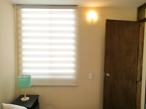 A room with a window covered by blinds, a bedside lamp, a wooden door, and a thermostat on the wall. - Aparta Hotel Tocancipa (Tocancipá)