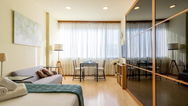 A hotel room with a bed, a sofa, a dining table with chairs, a TV, and a large mirror. - Holi-Rent Martín Villa (Sevilla)
