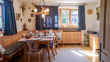 Un coin repas chaleureux avec une table en bois, des chaises et un banc. Il y a une fenêtre avec des rideaux, un lustre et une horloge au mur.