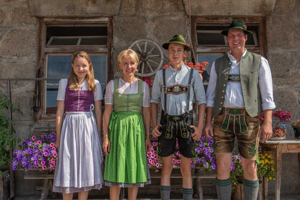 Four people in traditional attire standing in front of a stone building with a wheel and flowers.