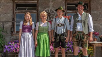 Four people in traditional attire standing in front of a stone building with a wheel and flowers.