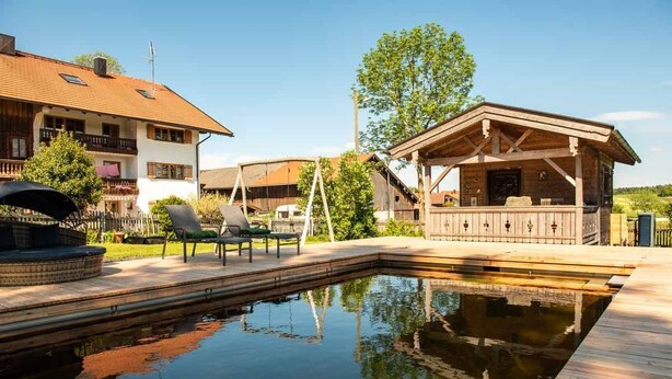 A swimming pool with a wooden deck, a covered seating area, and a building with a sloped roof.