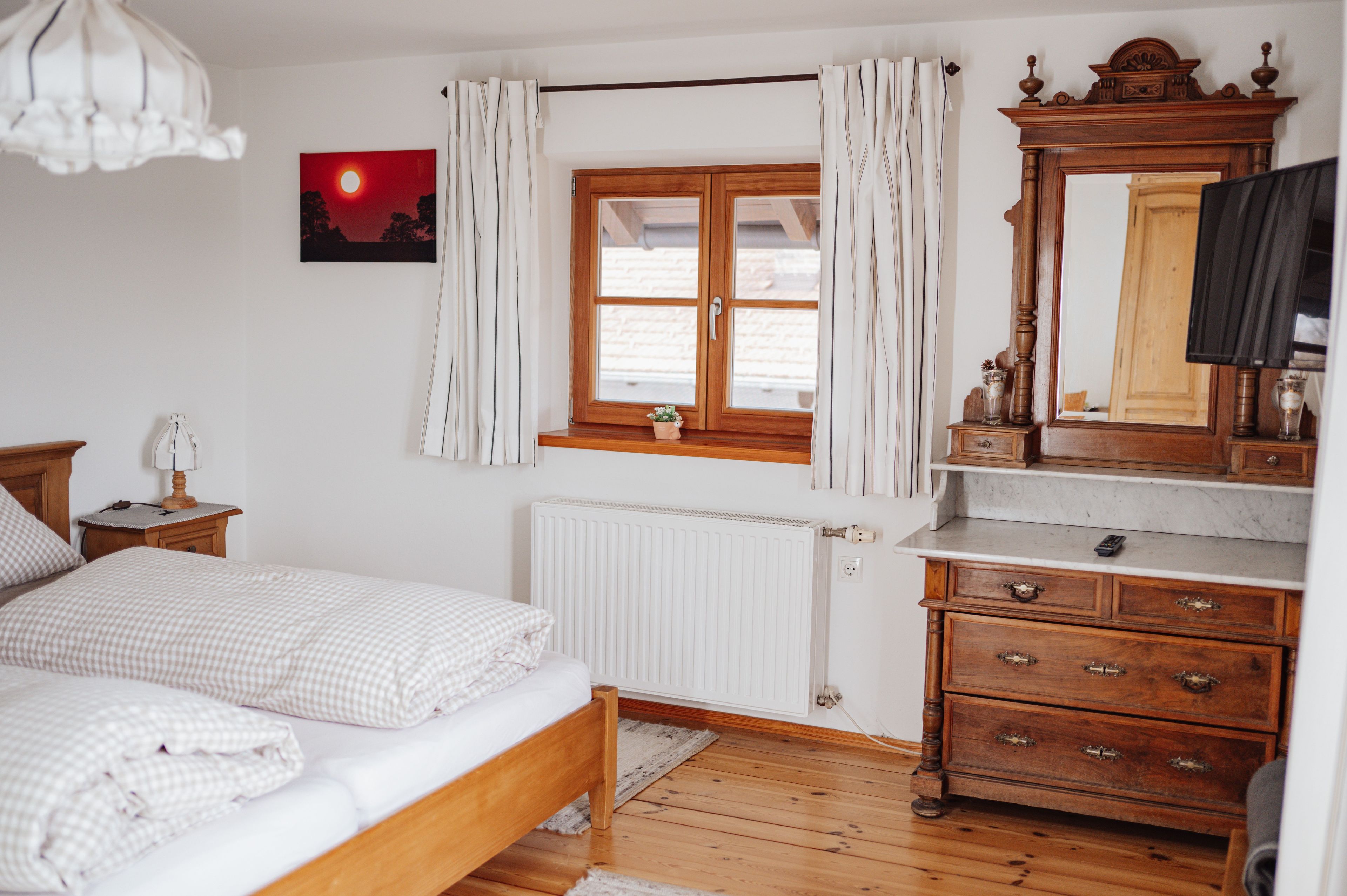 Une chambre à coucher comprenant un lit, une commode en bois, un miroir, une fenêtre avec des rideaux, un radiateur et un tableau accroché au mur.