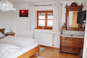 A bedroom with a bed, wooden dresser, mirror, window with curtains, radiator, and a wall-mounted picture. - Waldhauserhof - corner room (Waakirchen)