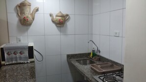 A kitchen with white tiled walls, a granite countertop, a sink, a stove, and two decorative teapots. - Kitnet one block from Pitangueiras beach (Guarujá)