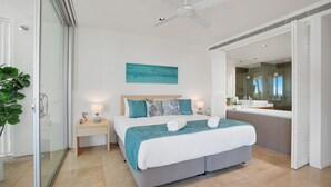 A modern hotel room with a large bed, a bedside table with a lamp, a small nightstand, a glass shower enclosure, and a view of the bathroom.