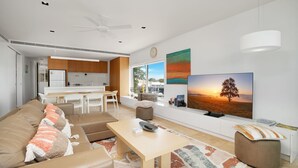 A modern living room with a sofa, coffee table, and TV. There's a kitchen area with wooden cabinets and a dining table.