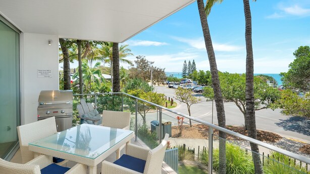 A balcony with a glass table and wicker chairs, overlooking a parking area and a view of the sea.