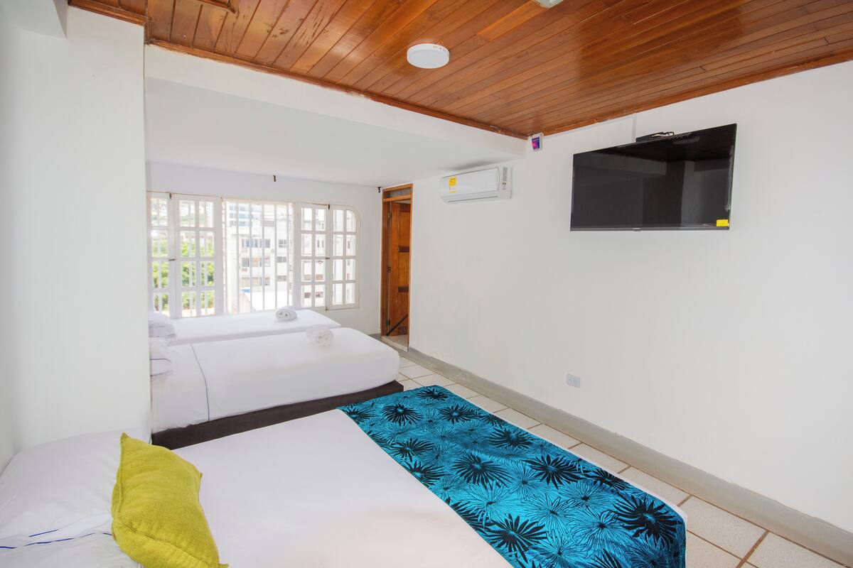 A hotel room with a wooden ceiling, a bed with a blue and white bedspread, a yellow pillow, and a TV mounted on the wall.
