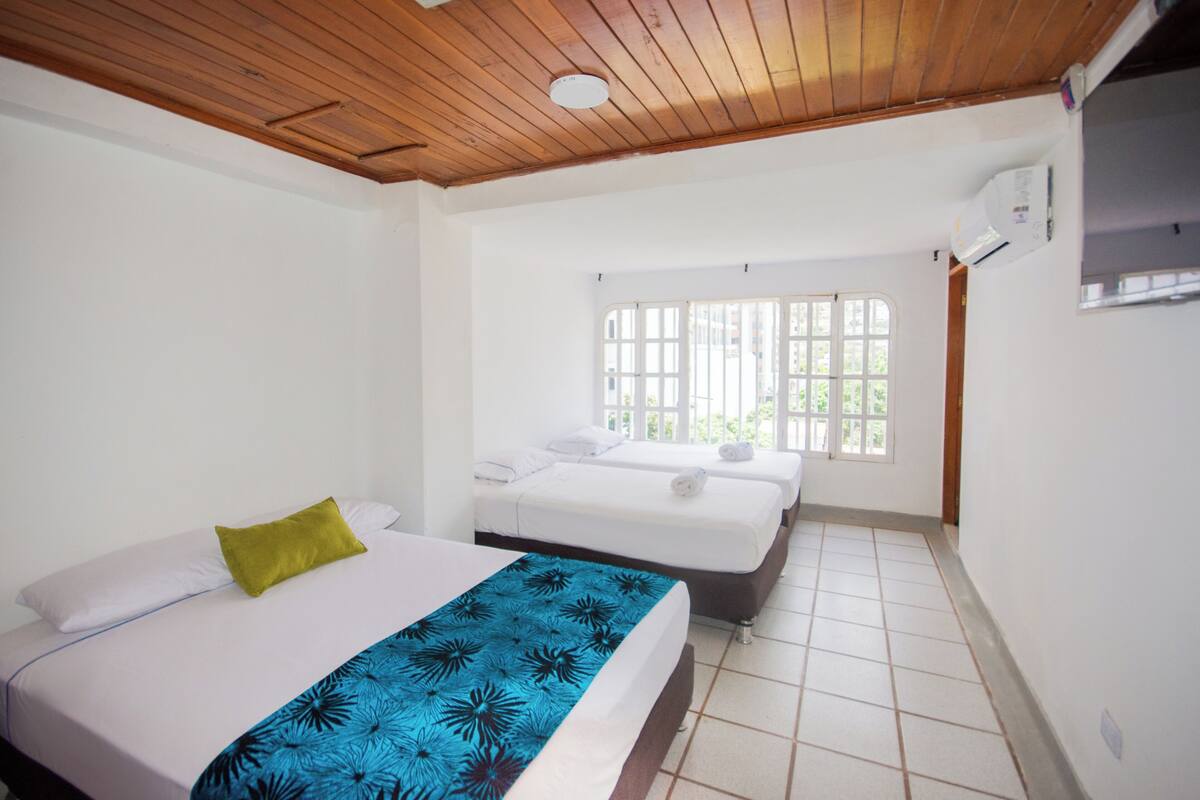 A hotel room with a wooden ceiling, a large window with bars, two beds, and a tiled floor.