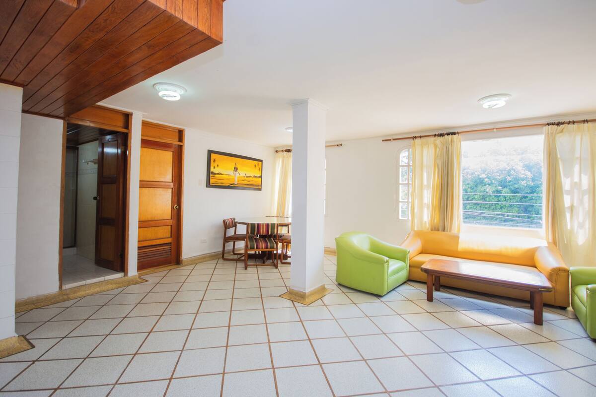 A spacious living area with a tiled floor, a green sofa, a wooden coffee table, and a dining table with chairs.