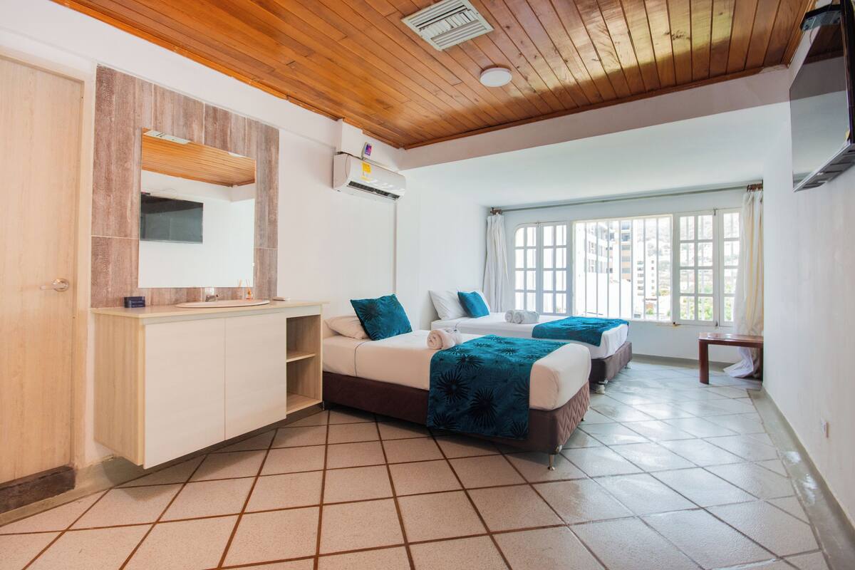 A hotel room with a wooden ceiling, a large window, a bed with a blue blanket, and a built-in wardrobe.