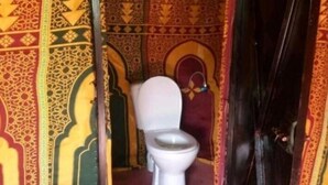 A small, narrow room with a toilet, a metal chair, and a patterned mat on the floor. - Dar Marhaba (Taouz)