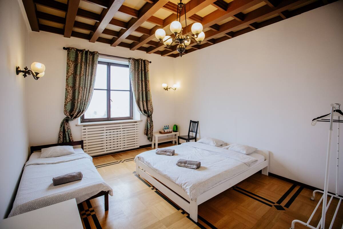 A bedroom with two beds, a window with curtains, a radiator, a chair, and a small table.