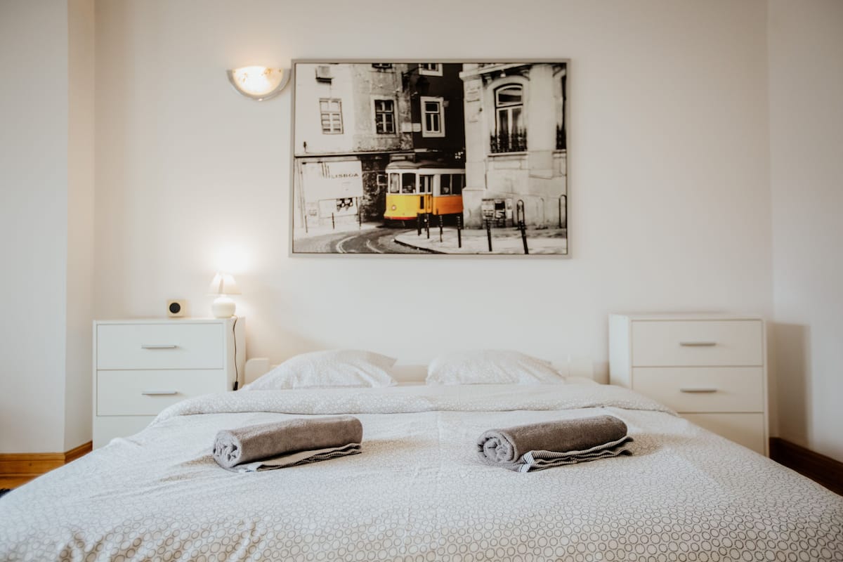 A hotel room with a bed, two bedside tables, a nightstand, a wall-mounted lamp, and a framed picture of a street scene.