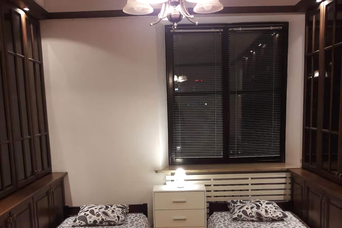 A bedroom with two beds, a chest of drawers, a window with blinds, and a ceiling light fixture.